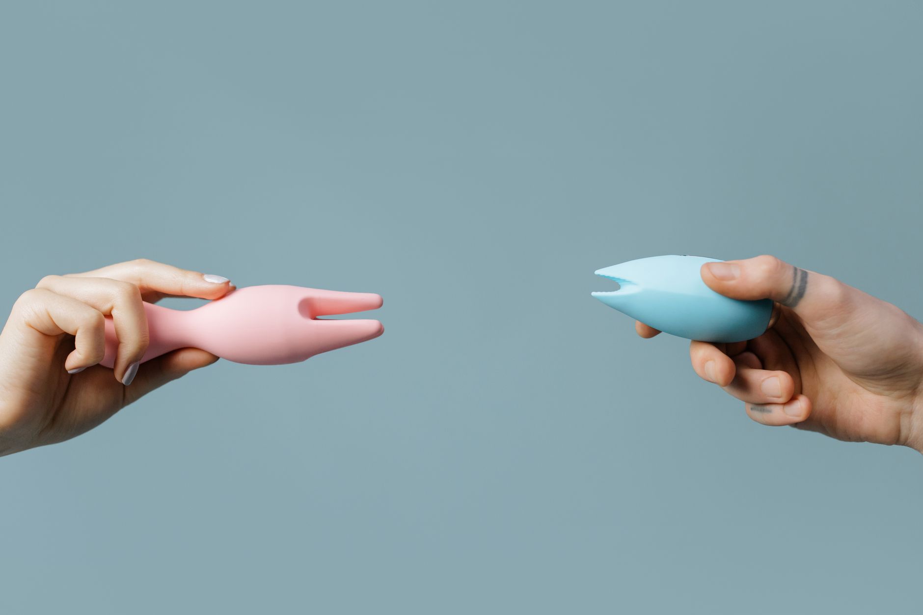Two hands holding pink and blue silicone vibrators, representing solo and partnered pleasure contexts