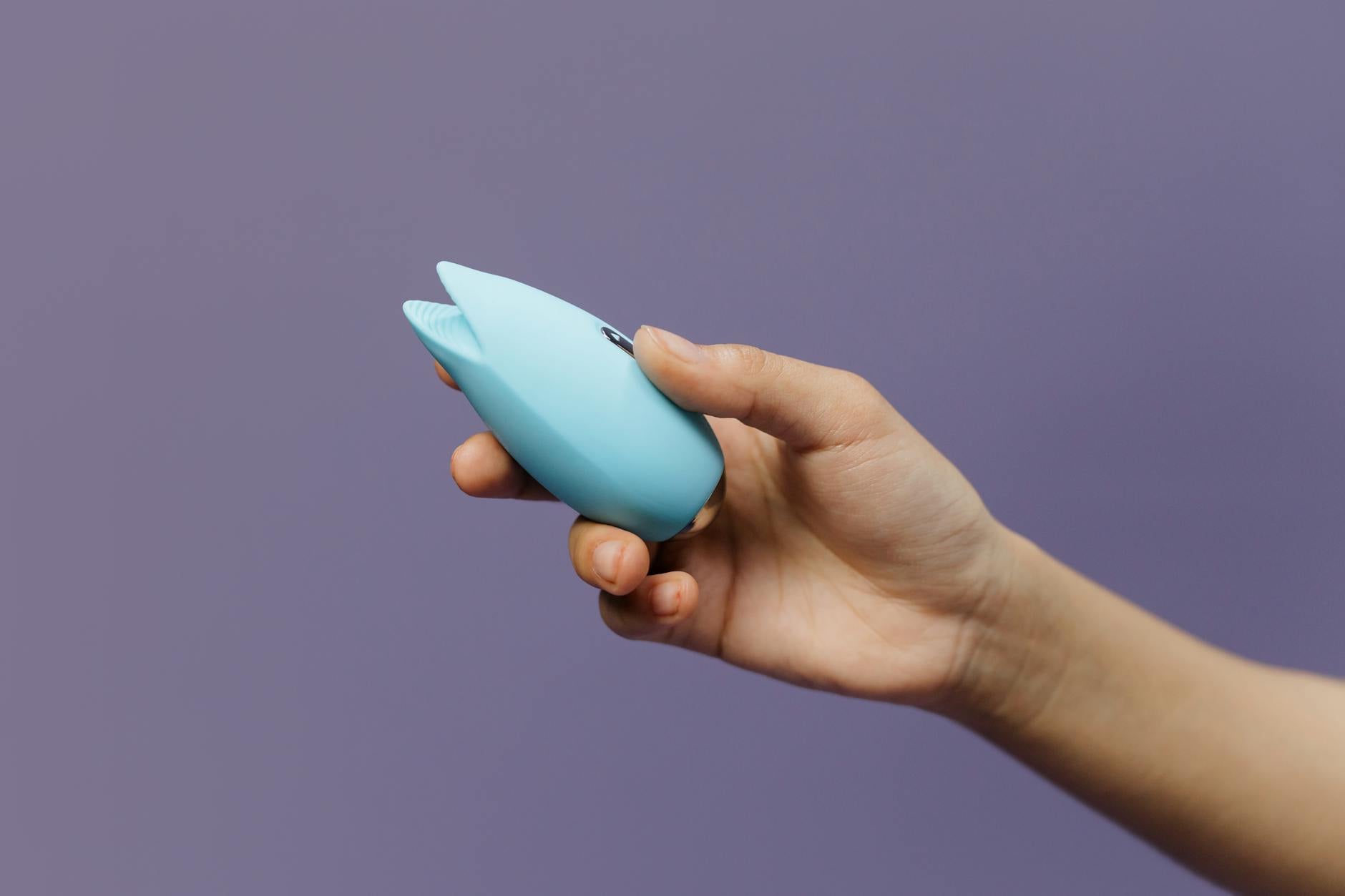 A blue silicone clitoral vibrator held gently in hand against a purple background, symbolizing pleasure recovery and self-care.